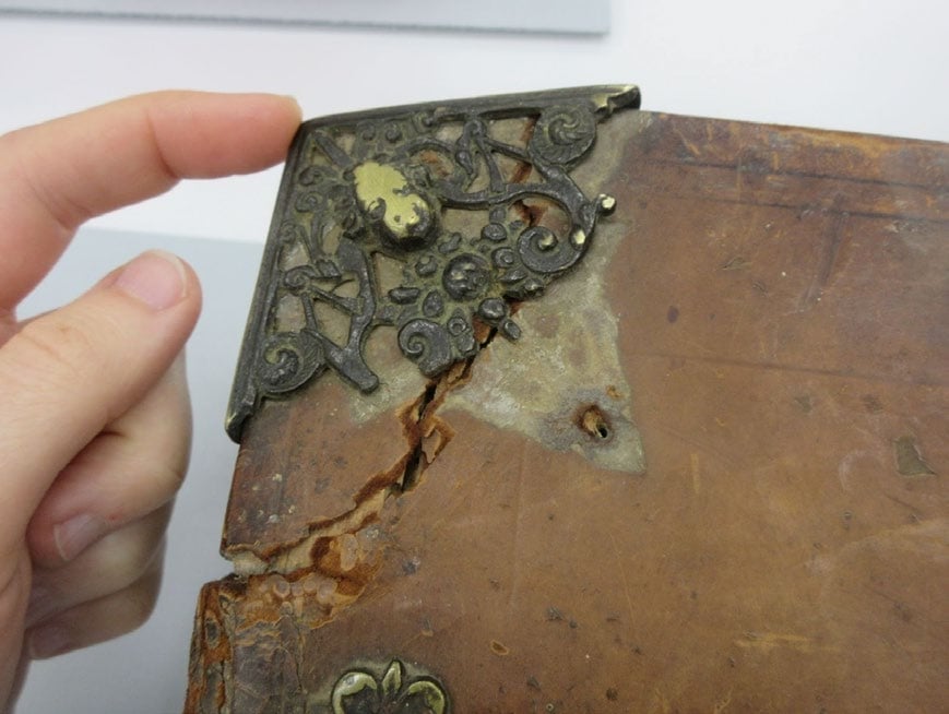 A hand holding the damaged corner of an old, leather-bound book with an ornate metal corner protector.