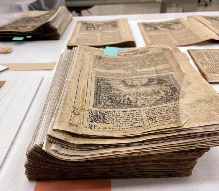 A stack of old, yellowed papers with printed text and illustrations sits on a table, surrounded by other similar documents.