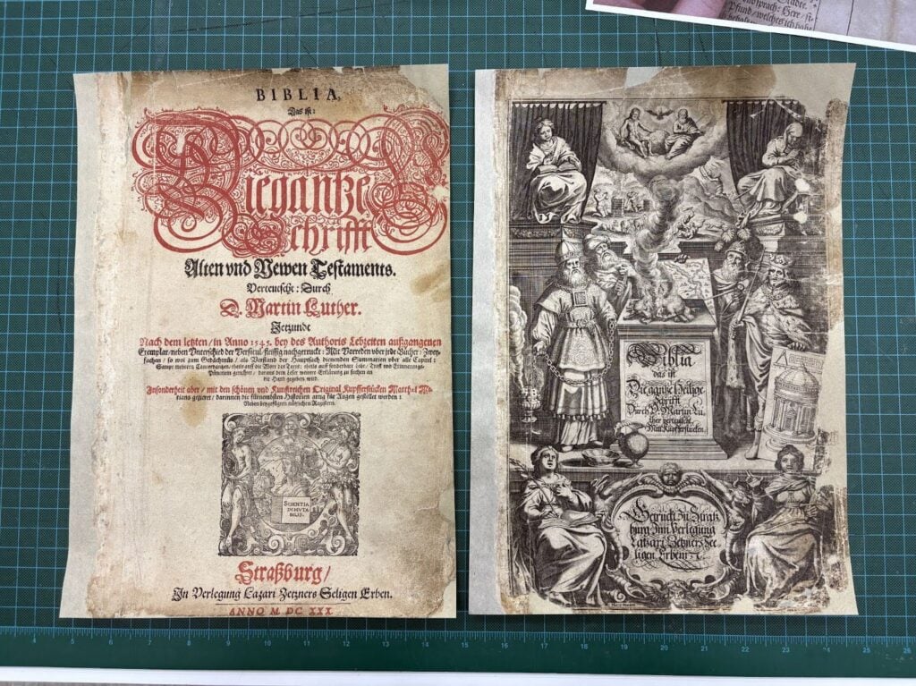 Two pages from an old German Bible, one with ornate red and black text, the other with detailed black-and-white religious illustrations. Both are displayed on a green cutting mat.