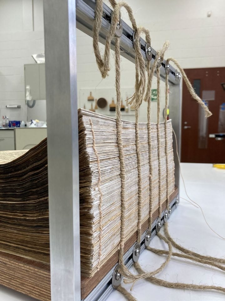 A stack of pages is held upright in a metal frame with multiple strings threaded through pulleys, likely for bookbinding or paper alignment in a workspace.