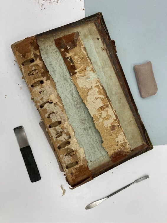 An old, damaged book cover with its spine missing is laid open on a table, alongside a metal tool, a rectangular eraser, and a utility knife.