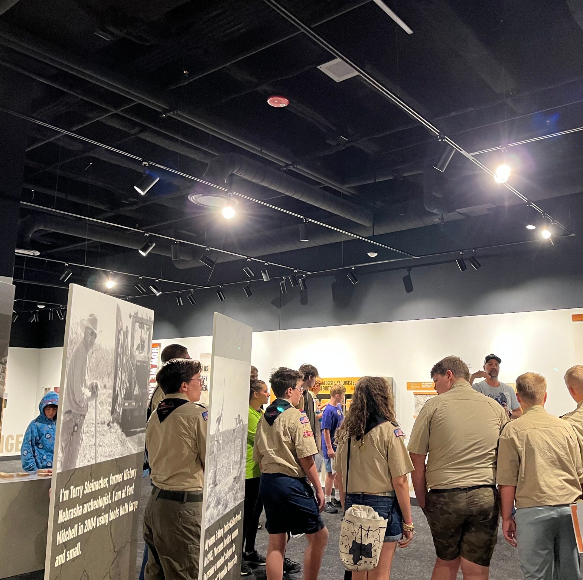 A group of people, mostly wearing tan uniforms, stand and look at exhibits inside a museum with overhead lighting.