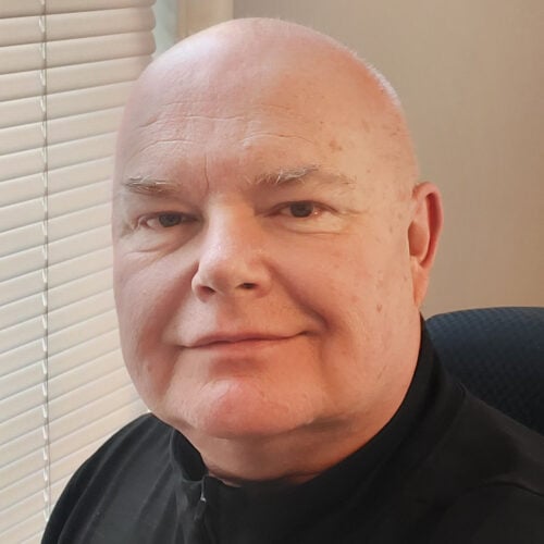 A bald man with light skin wearing a black shirt sits in front of a window with closed blinds, looking at the camera with a neutral expression.