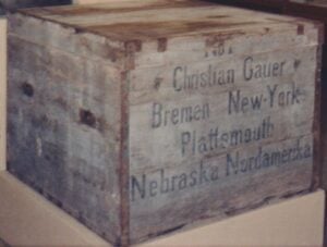 A large, weathered wooden shipping crate labeled with destinations including Bremen, New York, Plattsmouth, Nebraska, and Nordamerika, addressed to Christian Gauer.