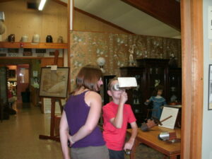 Two children stand in a museum room; one looks through a stereoscope viewer while the other stands beside watching. Various antiques and a doll are displayed in the background.
