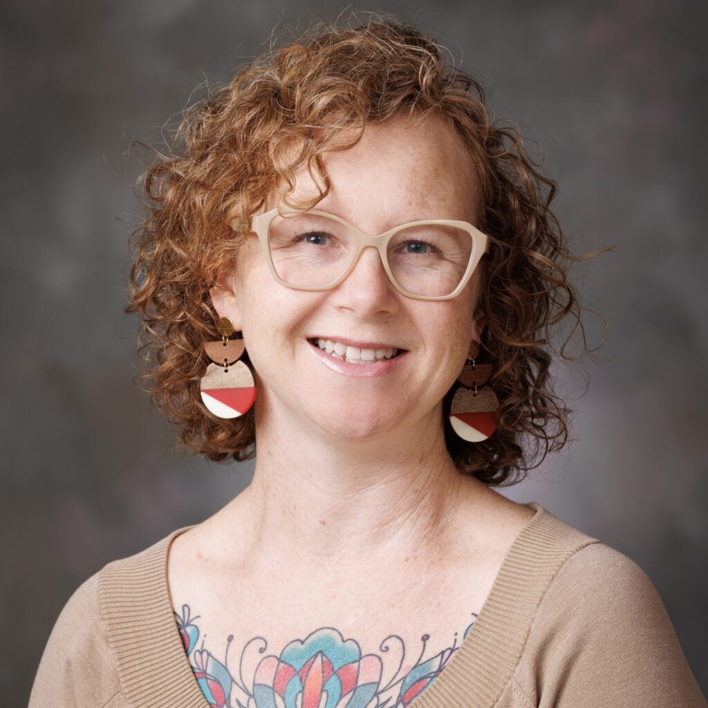 A person with curly red hair, beige glasses, large earrings, and a colorful floral chest tattoo smiles at the camera against a gray background.