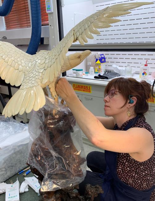 A woman wearing safety gear works on restoring a large bird sculpture in a laboratory setting, with tools and supplies visible nearby.