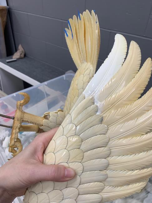 A hand holds a large artificial bird wing with cream-colored feathers; the wing appears to be part of a decorative or taxidermy piece in a plastic bin.