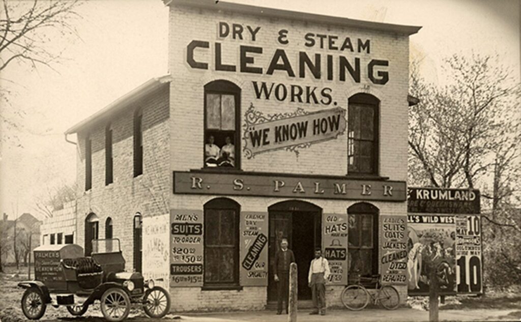 Two-story dry cleaning business building with large painted signs that say "Dry & Steam Cleaning Works. We Know How. R.S. Palmer. Men's suits to-order. $20.00 to $50.00. Trousers. $6.00 to &15.00. French dry and steam cleaning our speciality. Old hats made new. Hats blocked and primed $1.50. Ladies suits coats and jackets cleaning dryed and repaired." Three people stand outside, with a vintage car parked at the curb that has the following text on the side, "Palmer's Dry & Steam Cleaning...We Know How. Both Phones Bell 194. InD 321."