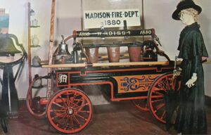 Antique fire wagon labeled "Madison Fire Dept. 1880" with buckets and equipment, displayed indoors next to a mannequin in period clothing.