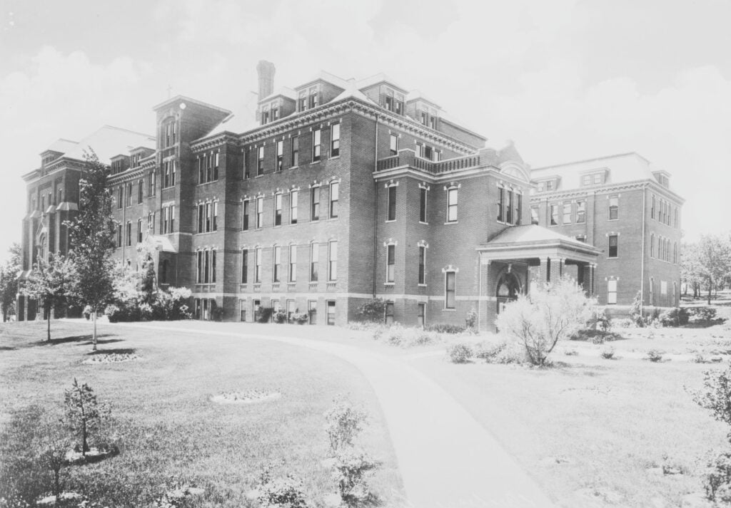 Black and white photo of a large, three-story brick building with multiple windows and a covered entrance, surrounded by a lawn and young trees.