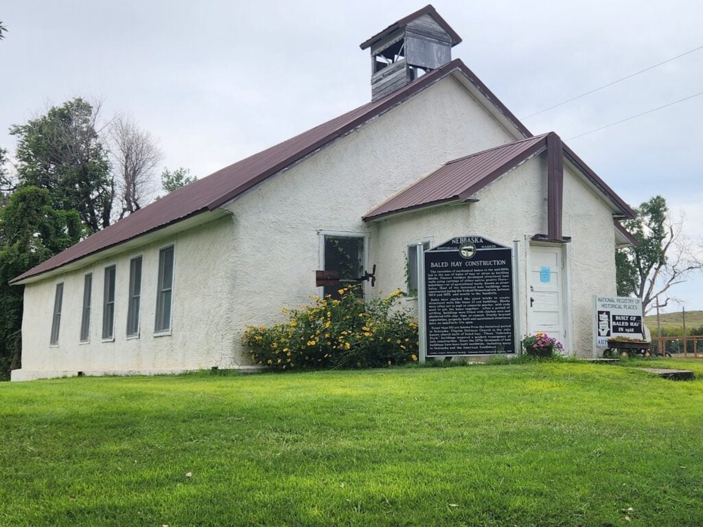 A small, white historic building with a brown roof, bell tower, and multiple signs and plaques on its lawn, surrounded by grass and flowers.