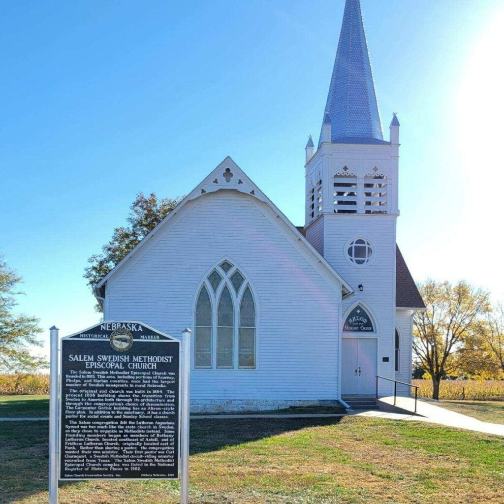 A white, wooden church with a tall steeple stands next to a historical marker sign that reads “Salem Swedish Methodist Episcopal Church” in Nebraska.