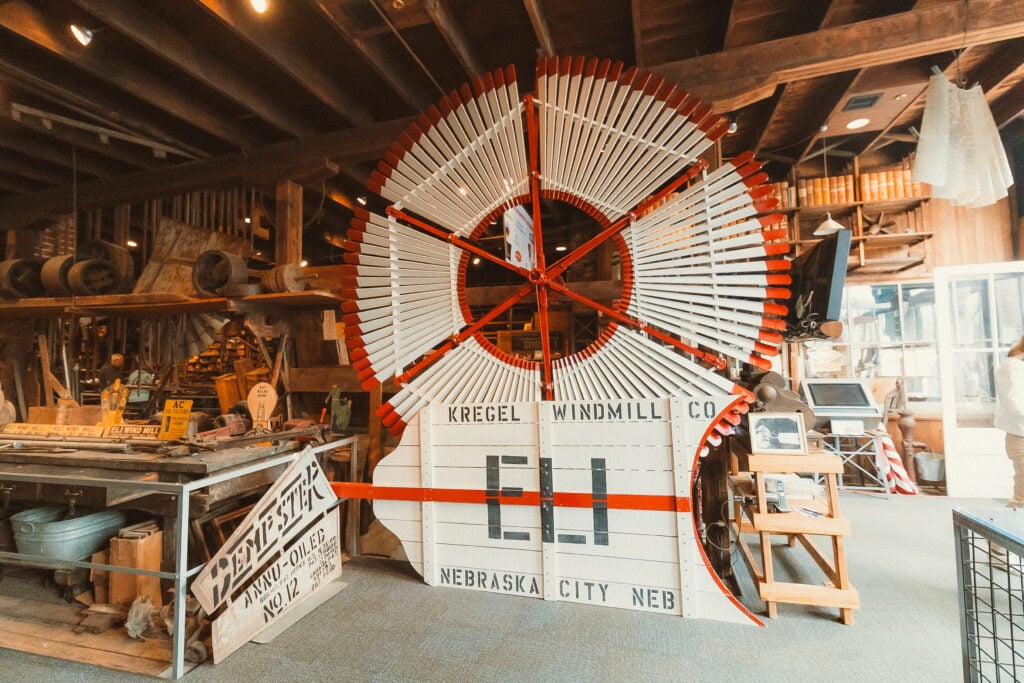 A large vintage windmill blade display marked "Kregel Windmill Co." and "Nebraska City, Neb." is inside a rustic workshop filled with tools, wood beams, and machinery.