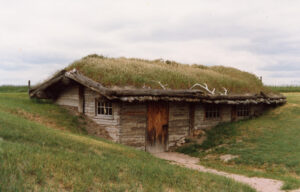 The Bordeaux Trading Post sod-roofed log cabin built into a grassy hillside, featuring a wooden door and three small windows under an overcast sky.