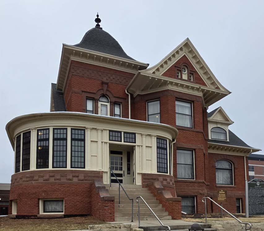 A historic red brick building with large bay windows, white trim, a turret, and a steeply pitched roof under a cloudy sky.