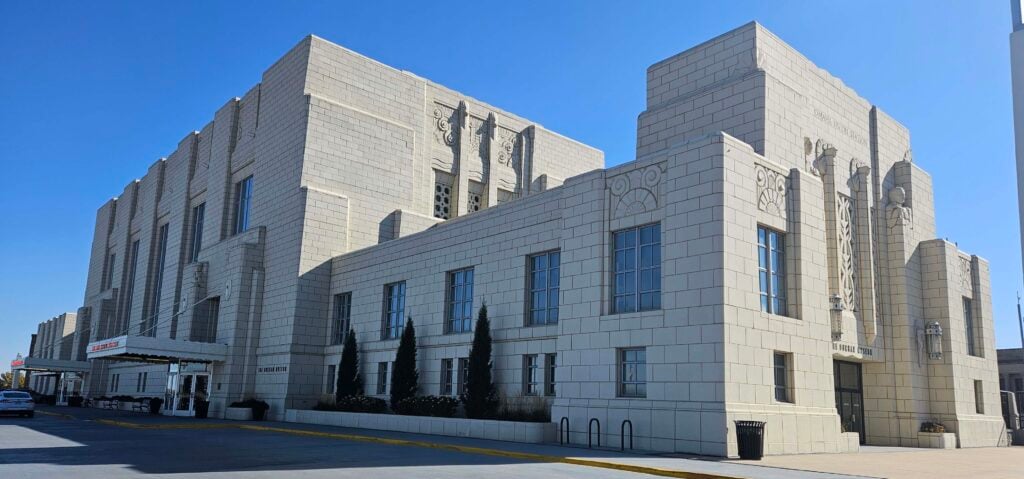 A large, light-colored Art Deco building with geometric designs, tall windows, and decorative elements stands under a clear blue sky.