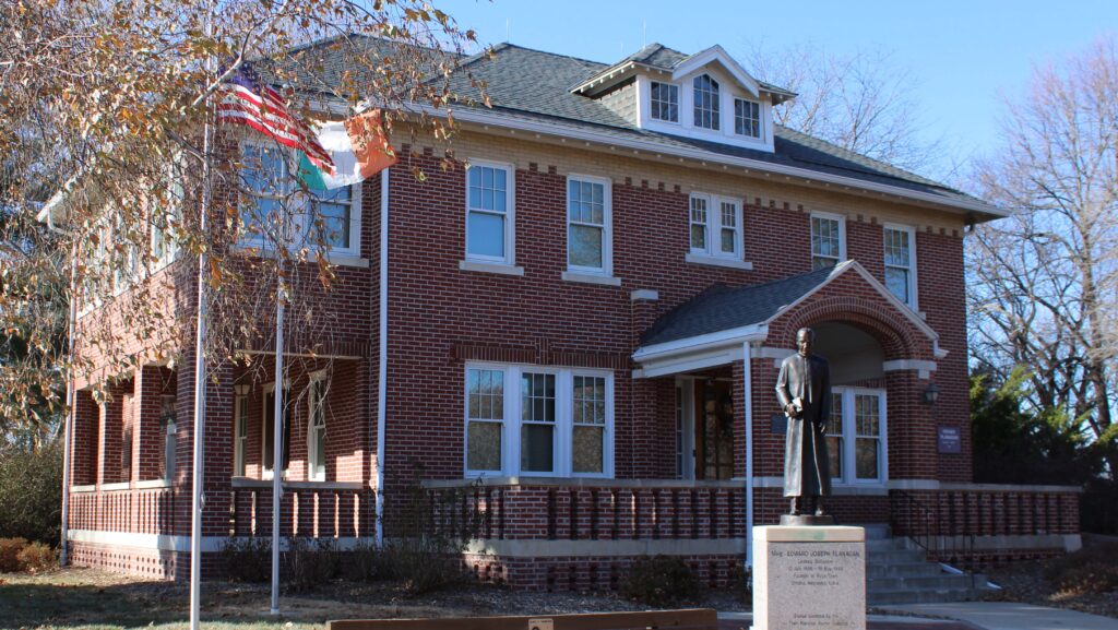 A two-story red brick building with white trim, an American and Irish flag, and a statue of Father Flanagan on a pedestal in front.