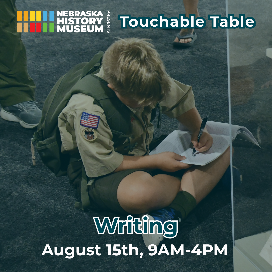 A boy in a scout uniform sits on the floor writing in a notebook at the Nebraska History Museum's "Touchable Table: Writing" event on August 15th, 9AM-4PM.