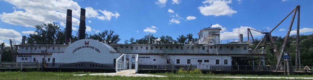 Side profile of the Captain Meriwether Lewis, as text says on the side of the ship which is docked. The ship has two smokestacks on one end and equipment on the other. A gated bridge to go aboard is attached to the side.