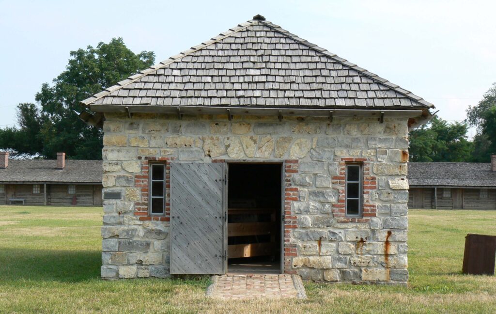 A small stone building with a wooden shingle roof and open wooden door stands in a grassy area, with larger stone buildings in the background.