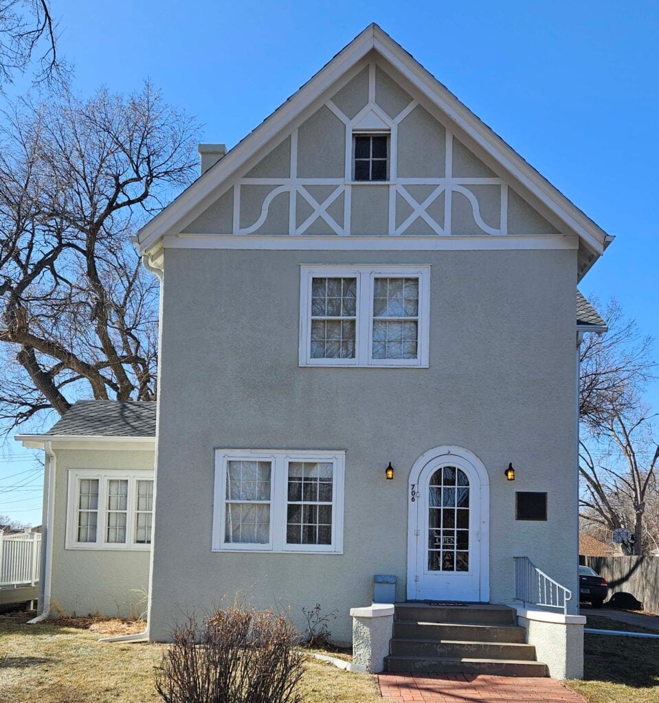 A two-story, light gray house with a steep gabled roof, white trim, and a small front porch with steps leading to an arched front door. Leafless trees and dry grass are visible.