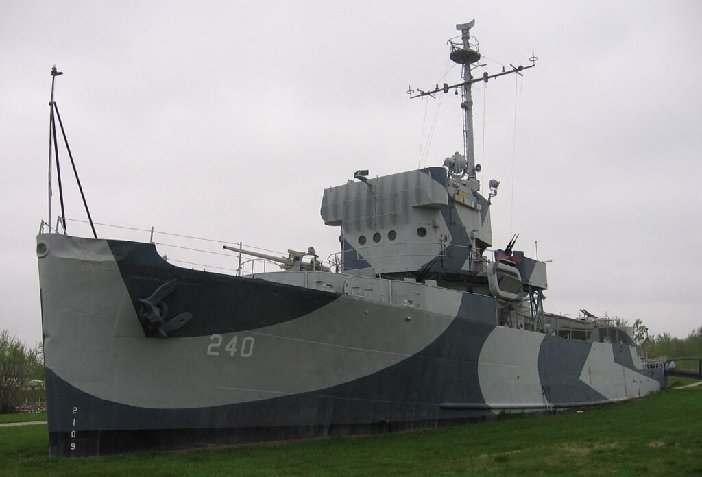 A decommissioned naval ship, number 240, painted in camouflage patterns, is displayed on a grassy field under an overcast sky.