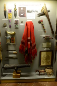 Museum display case with weapons, tools, historical artifacts, a red blanket, a model cannon and wagon, photographs, and framed documents labeled "Northern Monopoly.