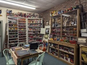 Room filled with shelves and cabinets displaying a large collection of toy trucks and tractors, with tables and chairs arranged for viewing books or documents.