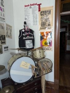 A vintage drum set is displayed in a room with Czech festival posters, a Czech polka band vest on a mannequin, and various framed photos and documents from Czech events on the wall.