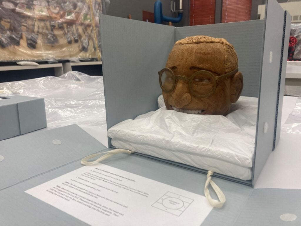 A carved coconut bust resembling Harry Truman with glasses displayed in a padded archival box, partially wrapped in white tissue paper, with instructions visible beside it.