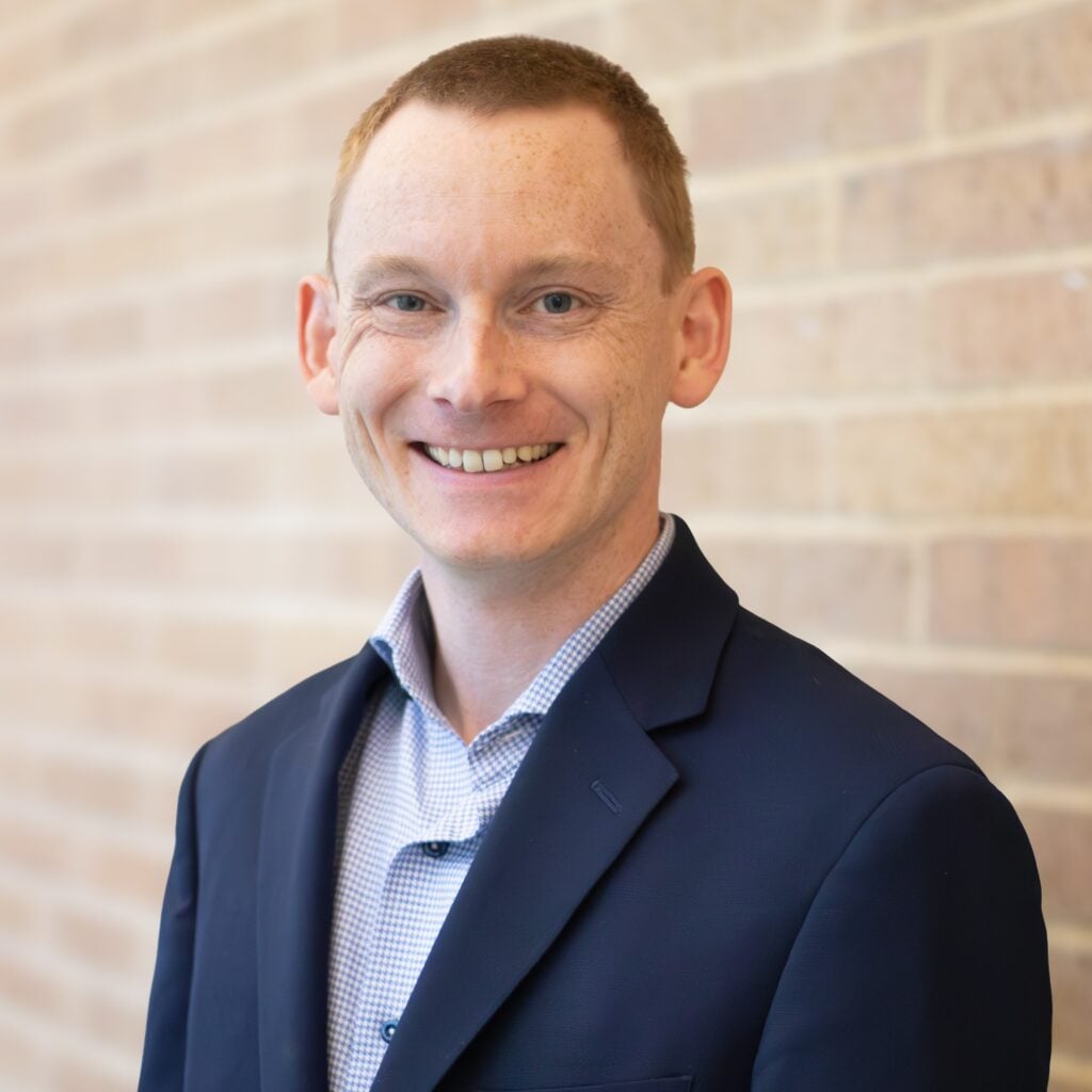 A man with short red hair wearing a dark blazer and checked shirt smiles while standing in front of a brick wall.