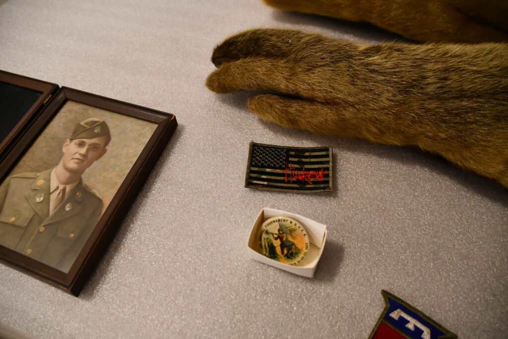 Framed photo of a soldier, a large animal paw, a black American flag pin, a circular pin, and a fabric patch displayed on a textured surface.