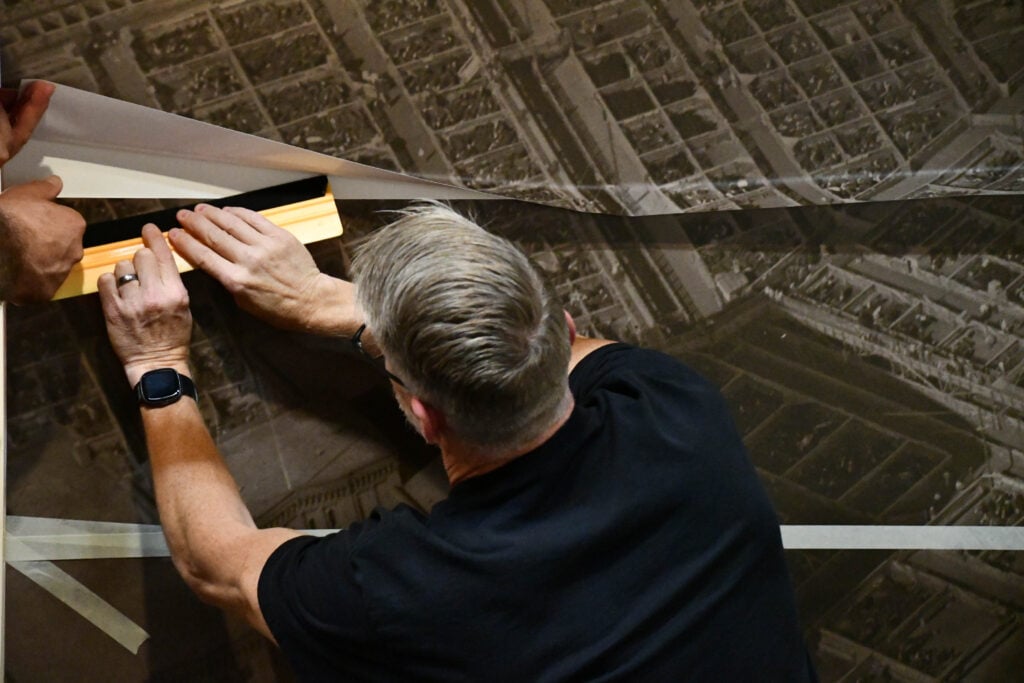 A person uses a tool to smooth or apply a large sheet of vinyl with a printed aerial image of the Omaha Stockyards onto a wall.