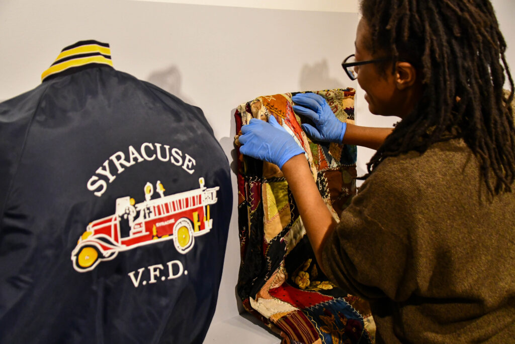 A person wearing blue gloves examines a colorful crazy quilt on a wall next to a Syracuse V.F.D. jacket.