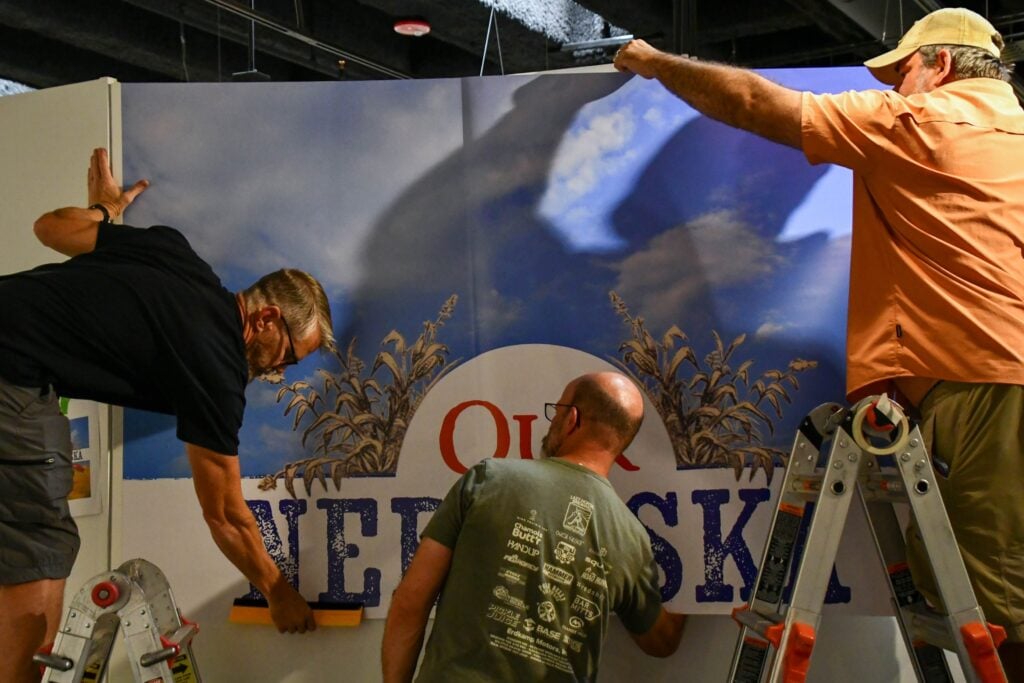 Three men install a large wall display with the word "Our NEBRASKA" partially visible, using ladders and tools under indoor lighting.