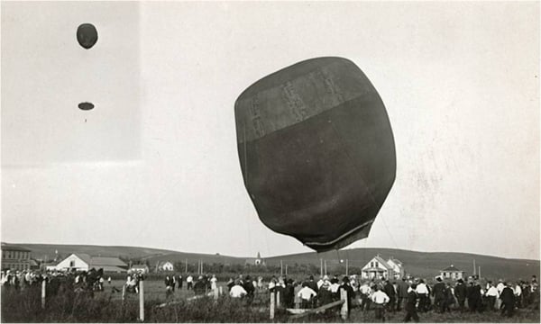 A large hot air balloon lifts off from a grassy field, surrounded by a crowd of people. An inset shows the balloon aloft with a man and parachute hanging from it.