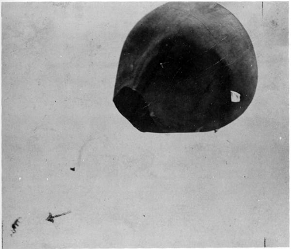 A large, dark, balloon-like object floats against a pale sky; a faint arrow points to a small detail in the lower left corner.