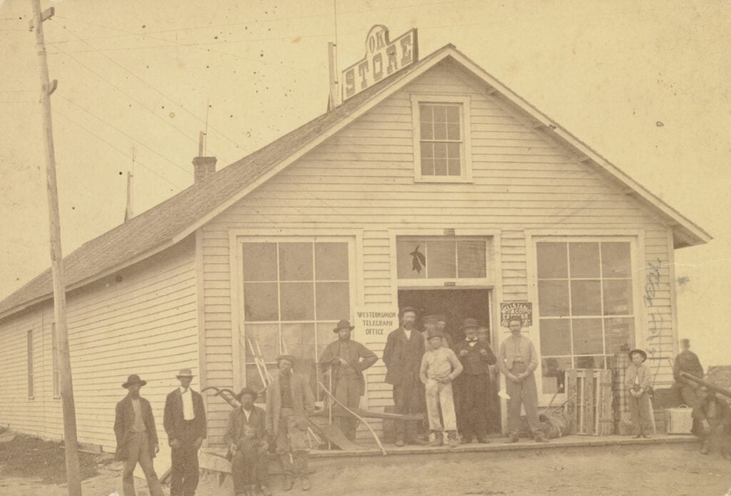 A sepia photo of people standing outside a wooden general store with large windows and a sign reading "Store" above the entrance. A “Western Union Telegraph Office” sign is posted on the building.
