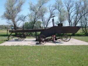 Large, old-fashioned mechanical grader with metal wheels and a long frame, displayed outdoors on a concrete pad with trees in the background.