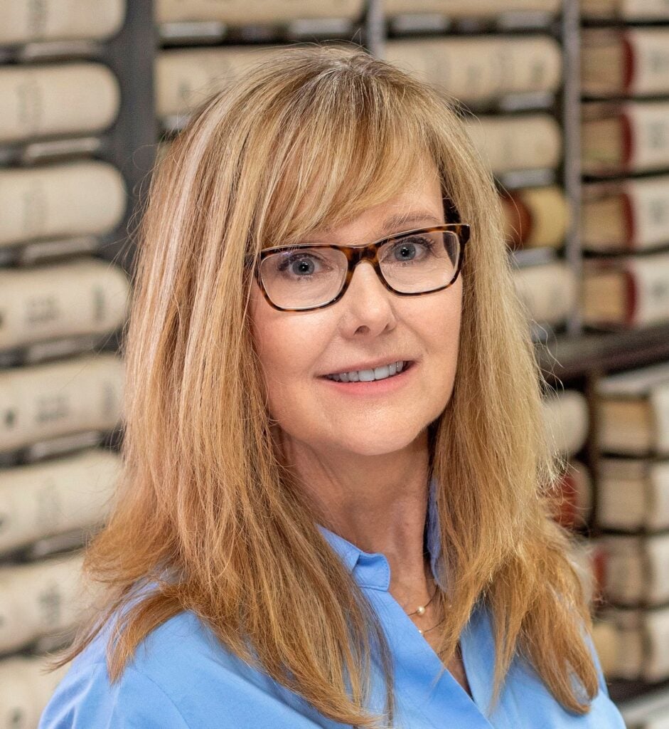 A woman with long blonde hair and glasses, wearing a blue blouse, is smiling in front of shelves filled with rolled documents.