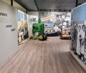 Exhibit with a green tractor, historical photos, and artifacts about Japanese Americans in Lincoln County, displayed in a museum setting with light wood flooring.