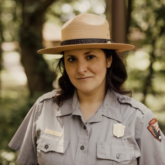 A park ranger in uniform and hat stands outdoors, looking at the camera with a neutral expression. Trees and greenery are visible in the background.