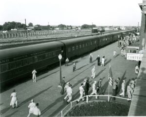 Black and white photo of people walking near a stopped passenger train beside the North Platte Canteen, with railroad tracks and buildings visible in the background.