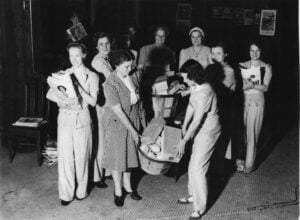 A group of women sort and collect stacks of newspapers and books into baskets in an indoor setting.