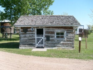 A small, old log cabin with a shingled roof, white picket gate, and one window sits on grassy land with a tree and water tower in the background.