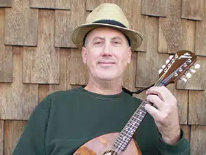 A man wearing a tan hat and dark green shirt holds a mandolin in front of a wooden shingle wall.