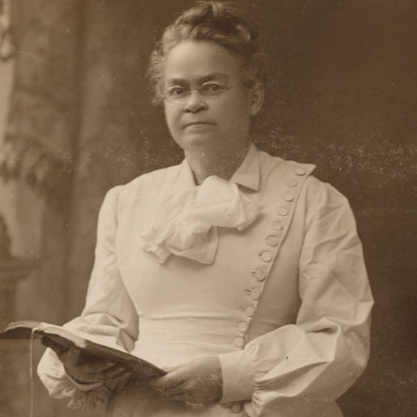 A woman wearing glasses and a white dress stands holding an open book, looking at the camera in a sepia-toned photograph.