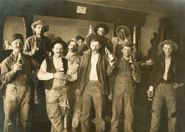 Nine men in work clothes and hats stand indoors, holding bottles and looking at the camera. The setting appears rustic and old-fashioned, possibly a bar or saloon.