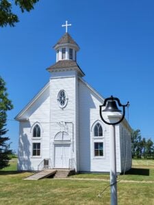 White wooden church with arched windows and a steeple; a metal bell on a post stands in the grassy yard under a clear blue sky.